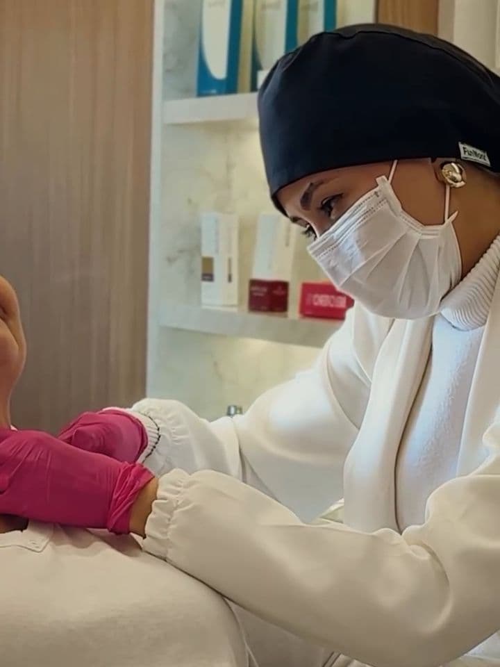 Juliane de touca e máscara atendendo uma paciente, com as prateleiras iluminadas ao fundo