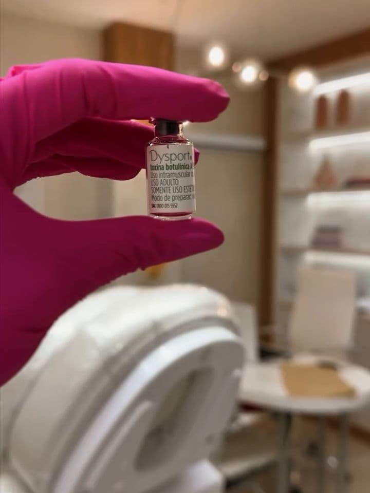 Luva magenta segurando um frasco, com o consultório ao fundo: cadeira de procedimento, luminárias e prateleiras iluminadas