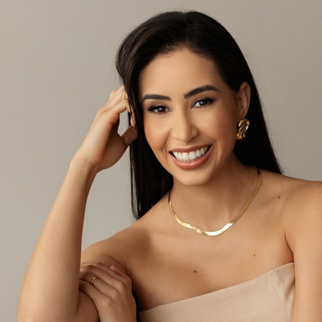 Juliane Mendanha sorrindo, de cabelo solto, colar e brincos dourados, diante de uma parede cinza